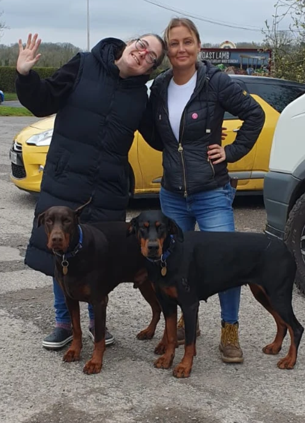 Lucy Humphrey e Katie James posam ao lado de dois cães doberman em uma área externa.